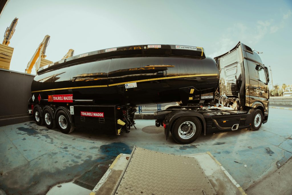 pexels-photo-16207913-16207913 Side view of a black tanker truck parked at an industrial shipping facility under clear skies.